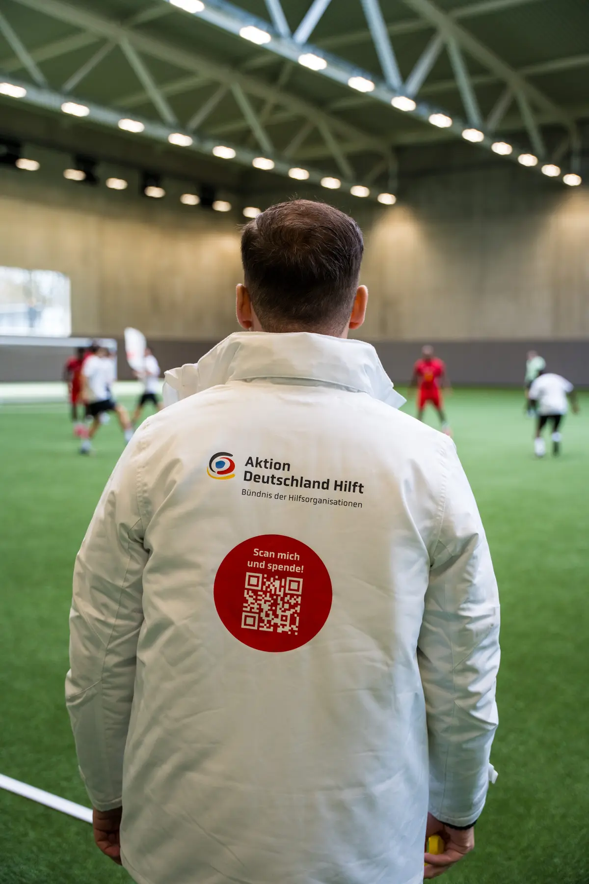 Helfer in Jacke von Aktion Deutschland Hilft mit Spenden-QR-Code am Spielfeldrand